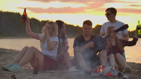 The-students-play-the-guitar-and-sing-songs-around-bonfire-on-the-beach-party.-This-is-soulful-summer-evening-with-the-best-friends-and-beer.